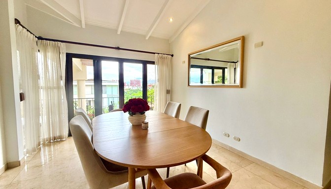Comedor moderno con mesa ovalada de madera, sillas tapizadas, centro de mesa con flores y decoración de mapas en la pared; al fondo se observa la sala de estar.